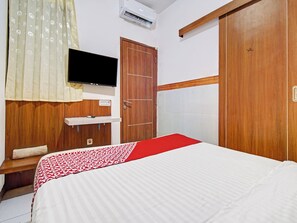 Habitación de hotel con cama, televisor montado en la pared, armario de madera y colcha estampada en rojo y blanco.