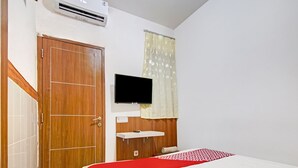 Un dormitorio con una puerta de madera, un televisor montado en la pared, una cama con colcha roja y blanca, y una ventana con cortina estampada.