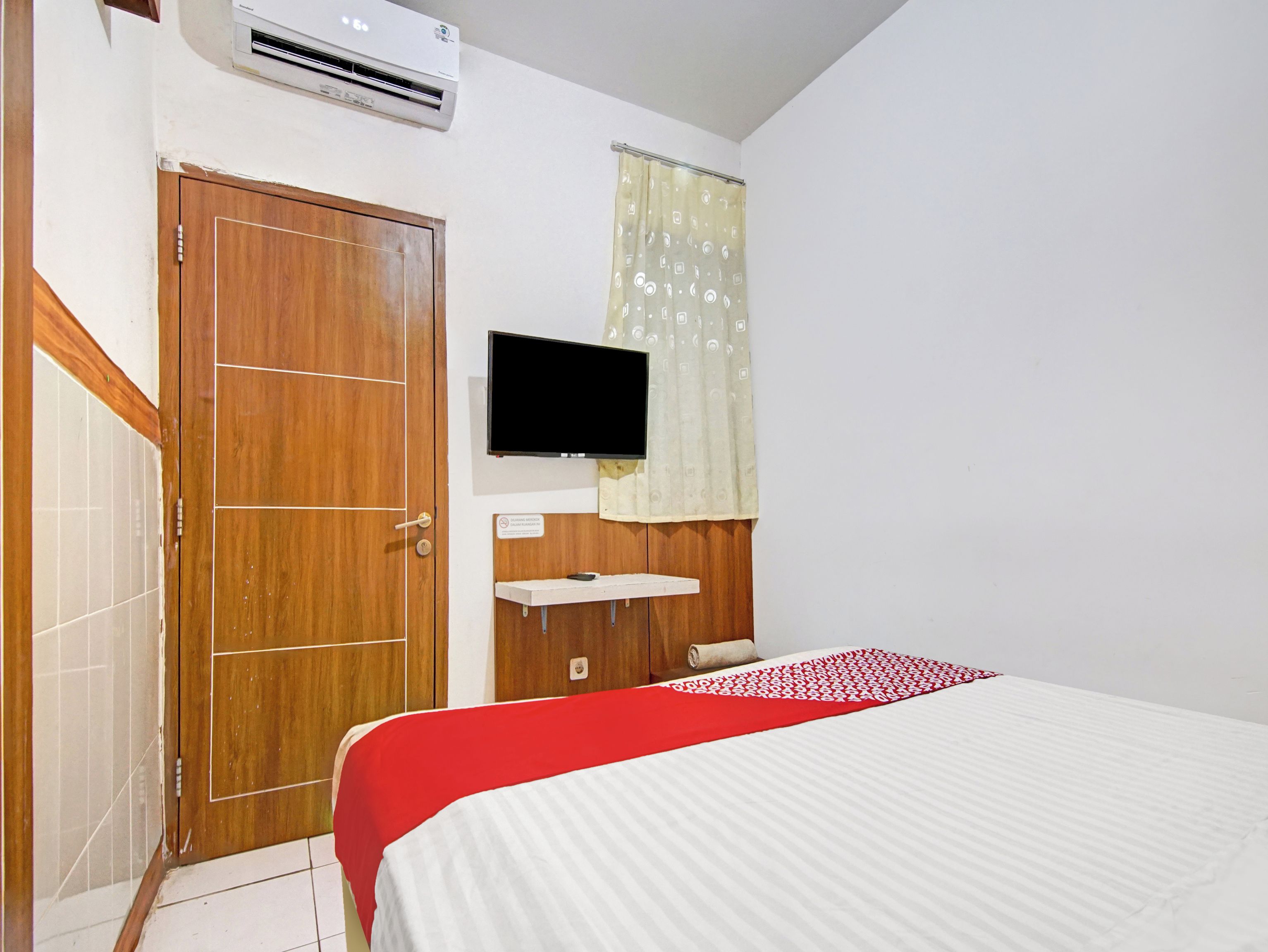 A bedroom with a wooden door, a television mounted on the wall, a bed with a red and white bedspread, and a window with a patterned curtain.
