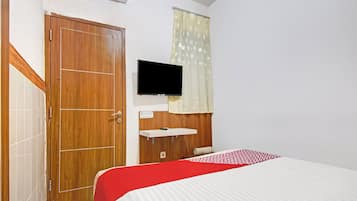 A bedroom with a wooden door, a television mounted on the wall, a bed with a red and white bedspread, and a window with a patterned curtain.