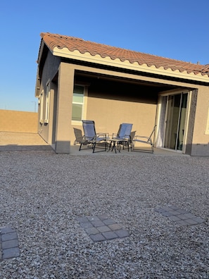 A building with a tiled roof, a covered patio area with two chairs and a table, and a gravel ground.