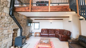 A spacious loft with a stone wall, a wooden staircase, a leather sofa, a wooden coffee table, and a fireplace. - Bowland Barn (Lancaster)