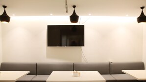 A modern interior with a long grey sofa, two white tables, and wooden chairs. A flat-screen TV is mounted on the wall above the sofa. - Hanting Hotel (Beijing)