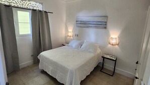 Een slaapkamer met een bed, nachtkastje, lamp, schilderij aan de muur en een raam met gordijnen.