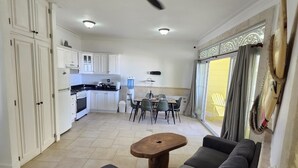 Een ruime woonkamer met een donkergrijze bank, een houten salontafel en een plafondventilator. Achtergrond toont een keuken met witte kastjes en een eethoek met een tafel en stoelen.