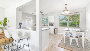 A modern kitchen with white cabinetry, a central island, and a dining area with white chairs and a table. - Cozy 2 BR Home, walking distance to Crystal Beach/Park - Quaint Family Getaway! (Palm Harbor)