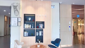Un área de recepción moderna con un mostrador de madera, dos sillas, una pequeña mesa con un florero y estanterías con varios objetos.