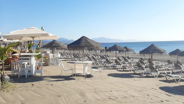 A beach with rows of sun loungers and thatched umbrellas, a table with chairs, and a view of the sea and mountains in the background. - 3 bedroom apartment in San Luis de Sabinillas (San Luis de Sabinillas)