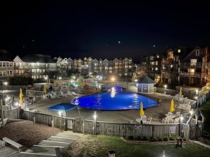 Vista noturna de um resort com uma piscina central, vários edifícios com varandas e áreas de estar ao ar livre.