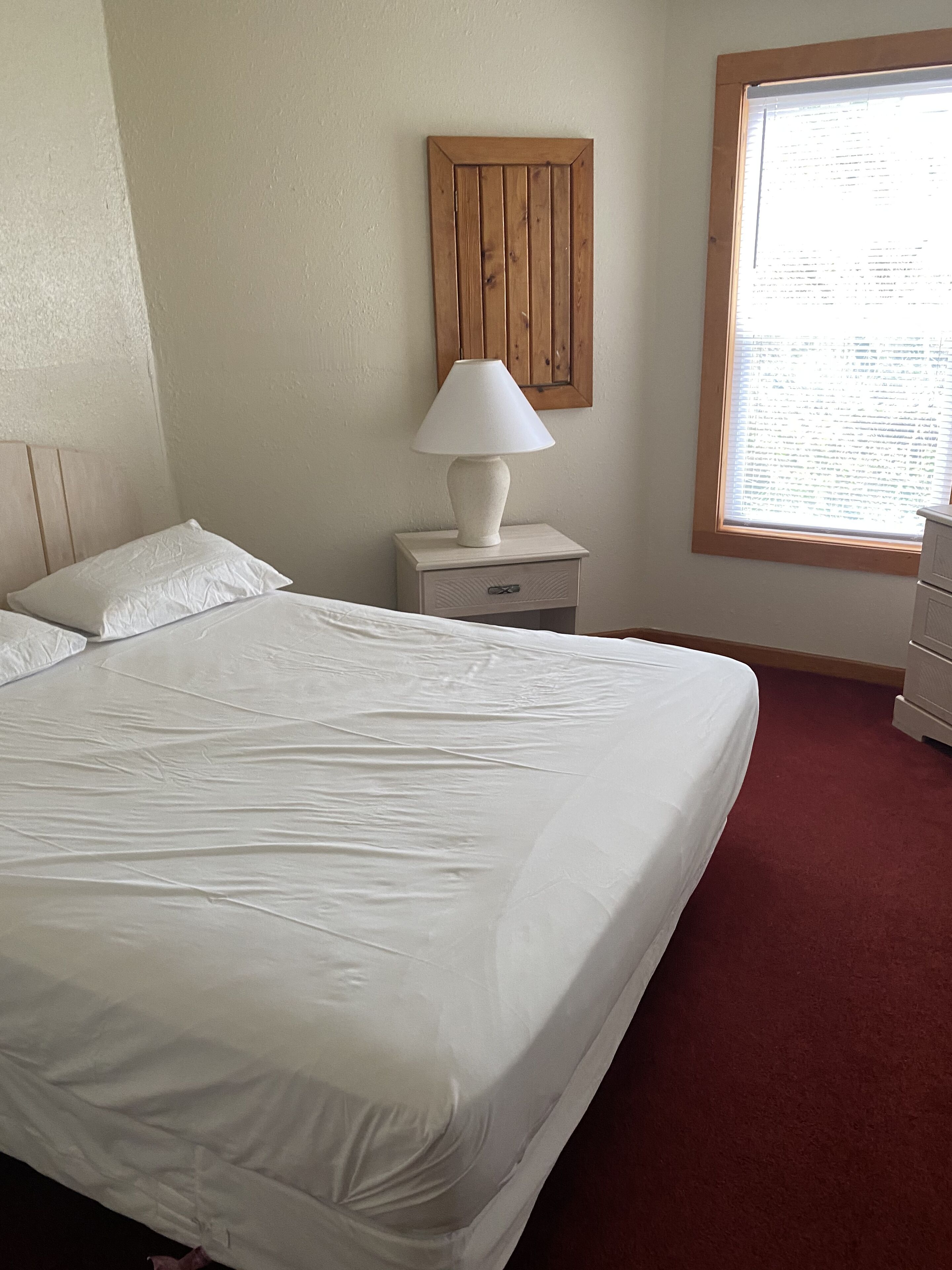 A neatly made bed with white linens, a bedside table with a lamp, and a window with blinds.