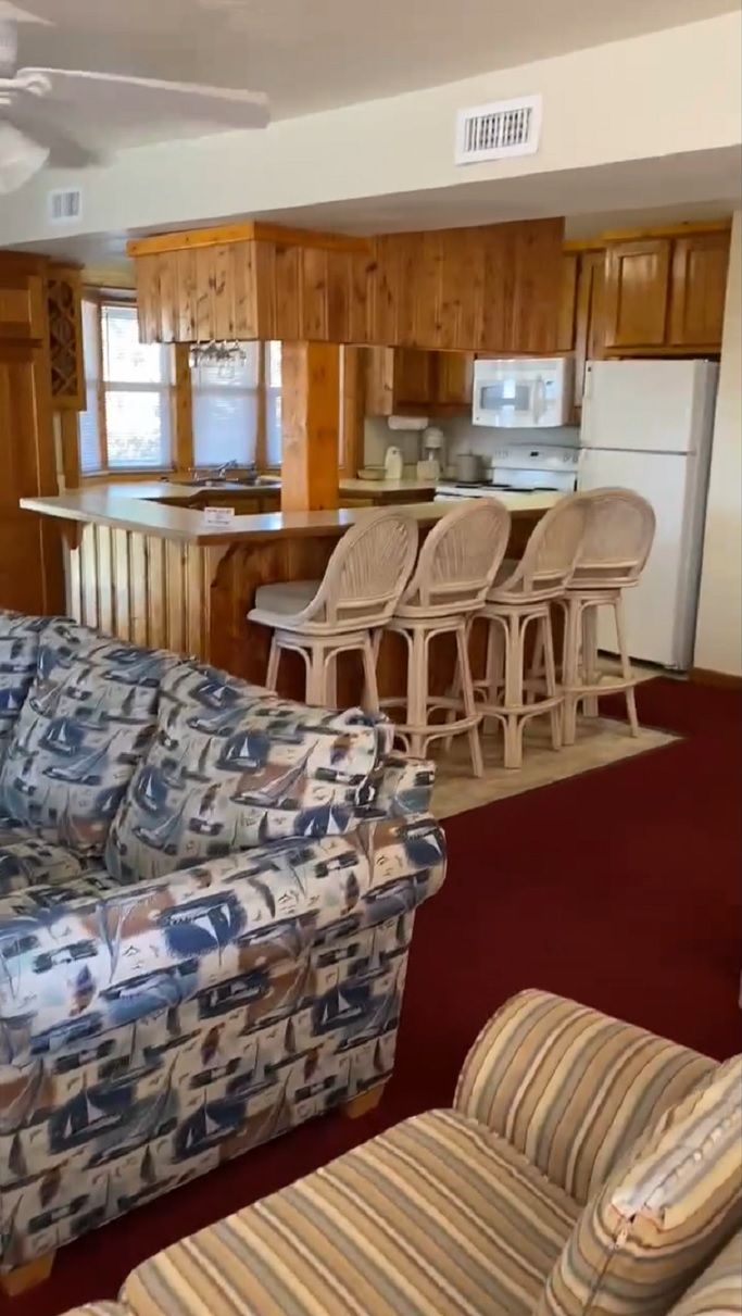 A living room with a patterned sofa and a striped chair, a kitchen with wooden cabinets and a microwave, and a ceiling fan.