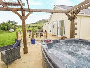 A hot tub with a wooden pergola, wicker furniture, and a view of a house and green landscape. - Cae Berllan (Pwllheli)