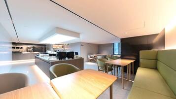 A modern hotel lobby with a kitchen area, seating arrangements, and a large window providing natural light.