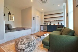 A living room with a sofa, ottoman, and a coffee table. There is a kitchen area with a microwave and cabinets.