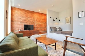 A living room with a brick wall, a flat-screen TV, a sofa, a coffee table, and a seating area with cushions.