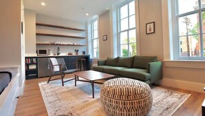 Una sala de estar moderna con un sofá verde, un reposapiés estampado, una mesa de centro de madera y una zona de cocina con microondas y estantes.