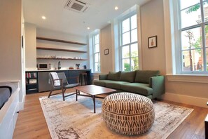 A modern living room with a green sofa, a patterned ottoman, a wooden coffee table, and a kitchen area with a microwave and shelves. - Prince Street Inn, Entire Building (Alexandria)
