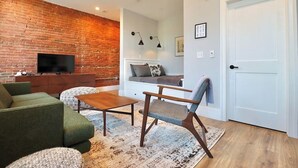 Una sala de estar con pared de ladrillo, televisor de pantalla plana, sofá verde, mesa de centro de madera y una alfombra estampada.
