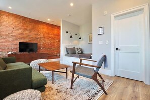 A living room with a brick wall, a flat-screen TV, green sofa, wooden coffee table, and a patterned rug. - Prince Street Inn, Entire Building (Alexandria)