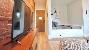 Una sala moderna con pared de ladrillo, televisor de pantalla plana, mesa de centro de madera, sofá blanco y un reposapiés estampado.