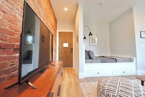 A modern living room with a brick wall, a flat-screen TV, a wooden coffee table, a white sofa, and a patterned ottoman.