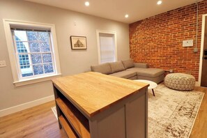 A living room with a wooden coffee table, a grey sofa, a patterned ottoman, and a brick wall. - The Old Town Suite at Prince Street Inn (Alexandria)