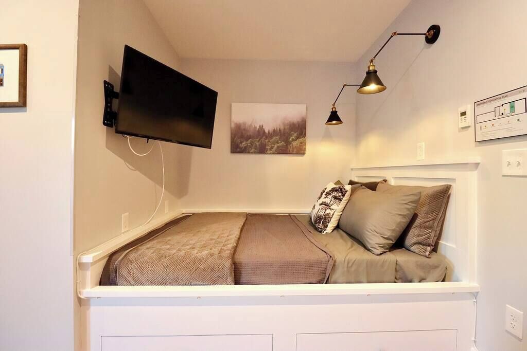 A small, cozy room with a bed, a mounted TV, a wall-mounted lamp, and a framed picture.