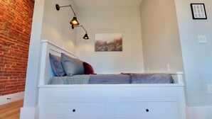 Ein Schlafzimmer im Loft-Stil mit schrägem Dach, einem weißen Bett mit Stauraum und einer Ziegelwand, beleuchtet durch eine hängende Leuchte.