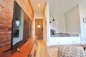 Una sala moderna con pared de ladrillo, televisor de pantalla plana, mesa de centro de madera, sofá blanco y un reposapiés estampado.