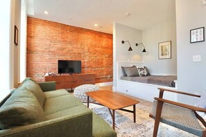 Una sala de estar con pared de ladrillo, un televisor de pantalla plana, un sofá, una mesa de centro y una zona de estar con cojines.