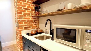 A kitchen with a brick wall, a stainless steel sink, a microwave, and wooden shelves above the counter. - The Prince Suite at Prince Street Inn (Alexandria)