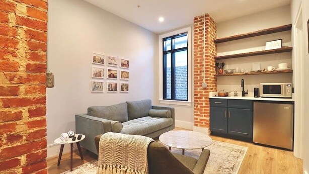 A compact living space with a sofa, a coffee table, a small side table, a brick accent wall, a kitchen area with a microwave, and a window with a view. - The Prince Suite at Prince Street Inn (Alexandria)