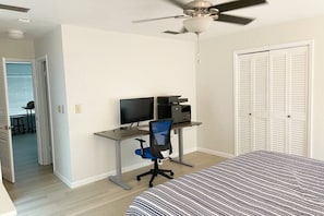 A bedroom with a bed, a desk with a computer and printer, a ceiling fan, and a closet.