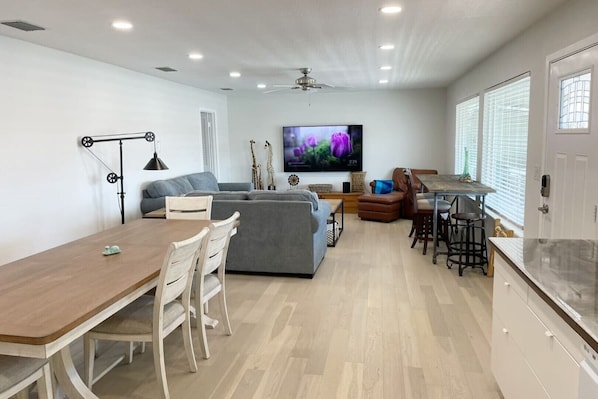 A modern living room with a dining area, grey sofas, a TV, and a kitchen with a large window. - Texas Grand Slam (City-by-the Sea)