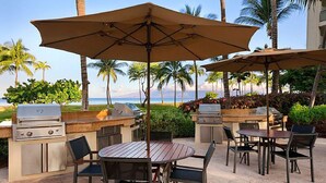 Ein Essbereich im Freien mit Grill und Sonnenschirm, mit Blick auf einen Strand mit Palmen und dem Meer.