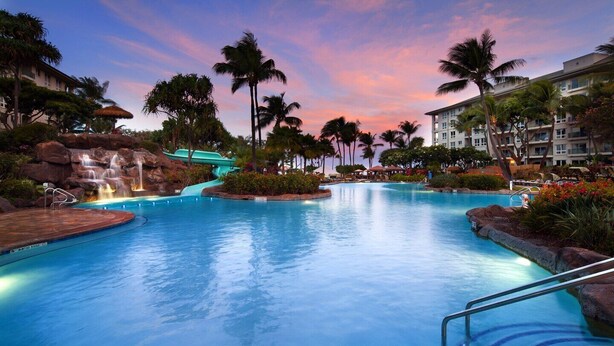 Ein großer, klarer Pool mit Wasserfall, umgeben von Palmen und einem Hotelgebäude.