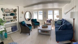 Ein gemütliches Wohnzimmer mit einem Korbssessel, einem blauen Sessel und einem passenden blauen Sofa. Ein Bücherregal mit Dekorationselementen und einem Seestern an der Wand.