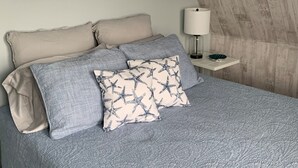 Ein Schlafzimmer mit Bett, Kissen, einem Nachttisch, einer Lampe und einem Wandbild.