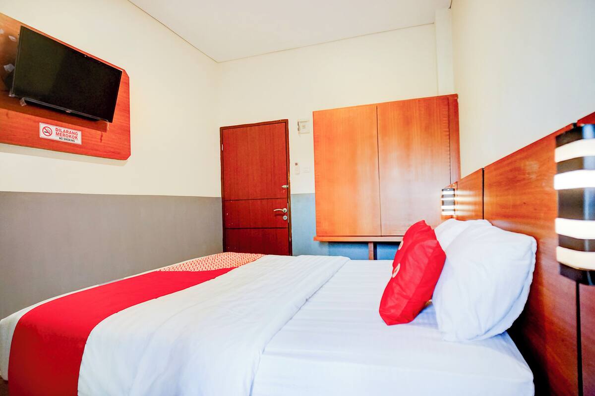 A hotel room with a bed, a wooden wardrobe, a television mounted on the wall, and a red and white bedspread.