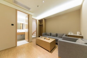 Una habitación de hotel moderna con suelo de madera, un sofá gris, una mesita de madera, una mesita de noche con lámpara y un armario con espejo.
