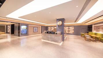 A modern hotel lobby with a reception desk, digital information kiosk, seating area, and a large digital display.