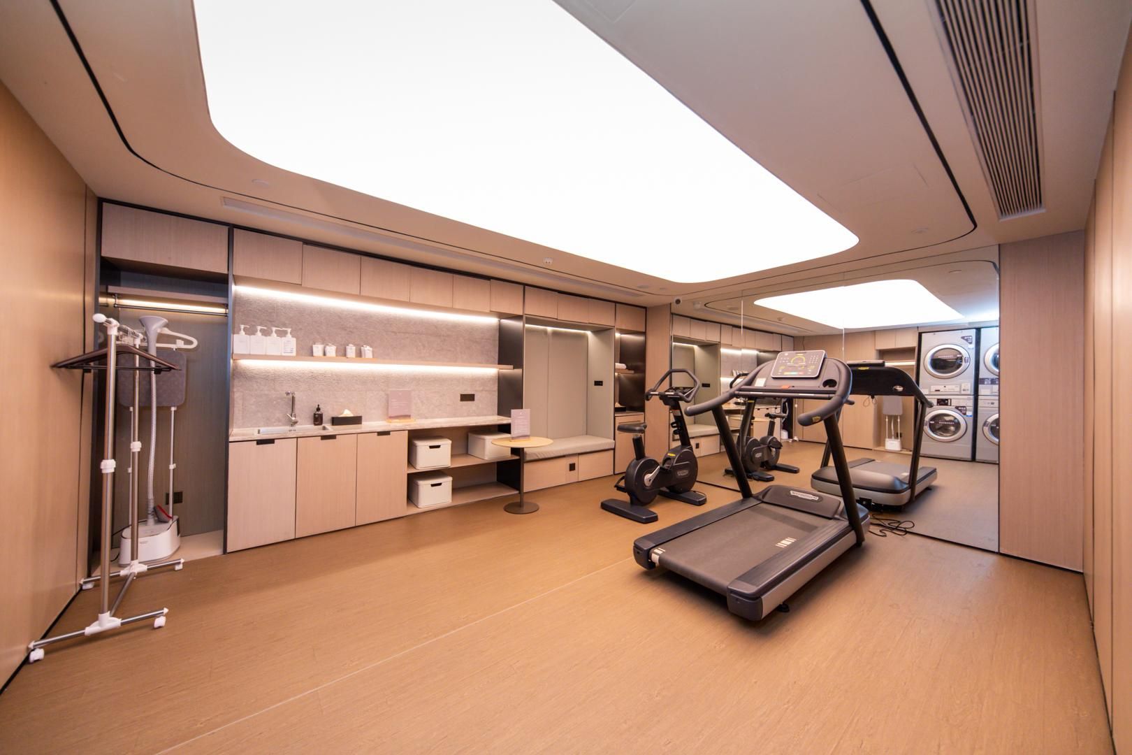 A modern hotel room with a large mirror, a treadmill, a stationary bike, and a compact kitchenette.