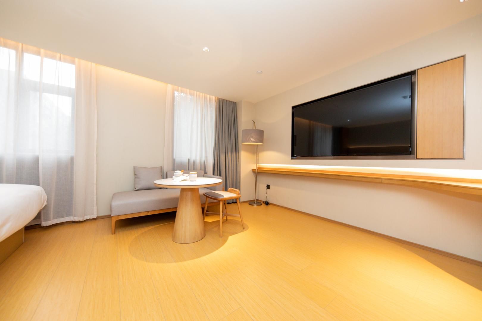 A hotel room with a bed, a round table with chairs, a sofa, a TV mounted on the wall, and a window with curtains.