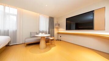 A hotel room with a bed, a round table with chairs, a sofa, a TV mounted on the wall, and a window with curtains.