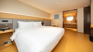 A modern hotel room with a large bed, bedside tables, a small table, a round mirror, and a bathroom with a sink and mirror.