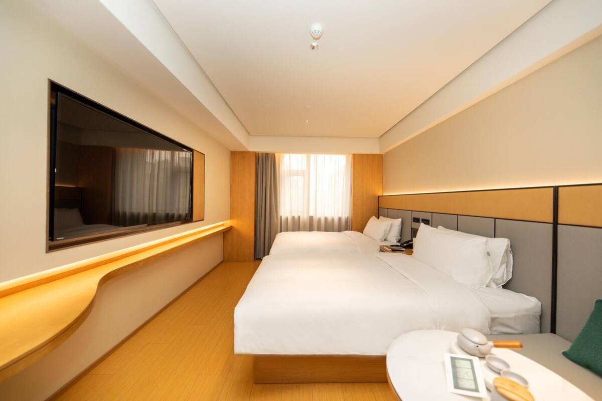 A hotel room with a large bed, a bedside table with a lamp, a TV mounted on the wall, and a window with curtains.
