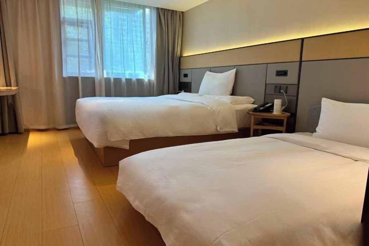 A hotel room with two beds, a bedside table with a lamp, and a window with curtains.