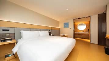 A modern hotel room with a large bed, bedside tables, a small table, a round mirror, and a bathroom with a sink and mirror.