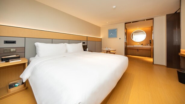 A modern hotel room with a large bed, bedside tables, a small table, a round mirror, and a bathroom with a sink and mirror. - Ji Hotel (Xi'an)