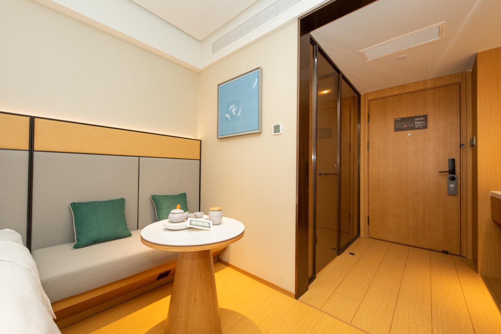 A hotel room with a sofa, a small round table with a tea set, a wall-mounted artwork, and a door leading to a hallway.